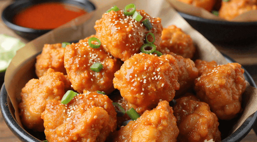 Bowl of Korean chicken bites glazed in spicy gochujang sauce, garnished with sesame seeds and green onions, with a dipping sauce in the background.