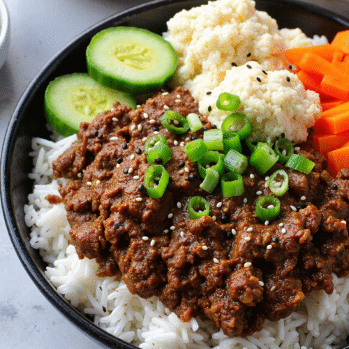 Korean ground beef bowl with rice, sliced cucumbers, pickled carrots, and cauliflower rice, topped with sesame seeds and green onions.