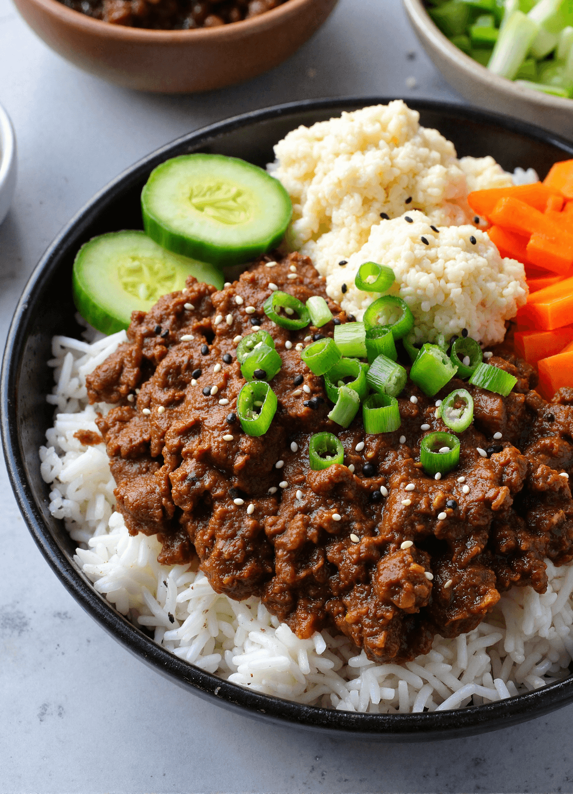 Korean ground beef bowl with rice, sliced cucumbers, pickled carrots, and cauliflower rice, topped with sesame seeds and green onions.