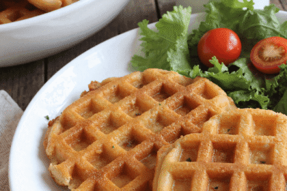 Plate of three golden crispy low carb hashbrown chaffles with a side of leafy greens and cherry tomatoes, perfect for healthy meal prep or low carb brunch.