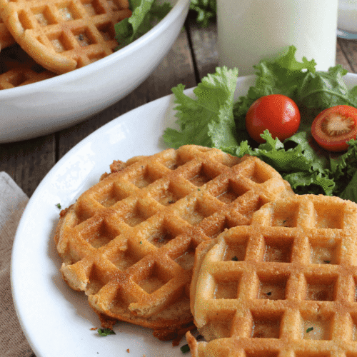 Plate of three golden crispy low carb hashbrown chaffles with a side of leafy greens and cherry tomatoes, perfect for healthy meal prep or low carb brunch.