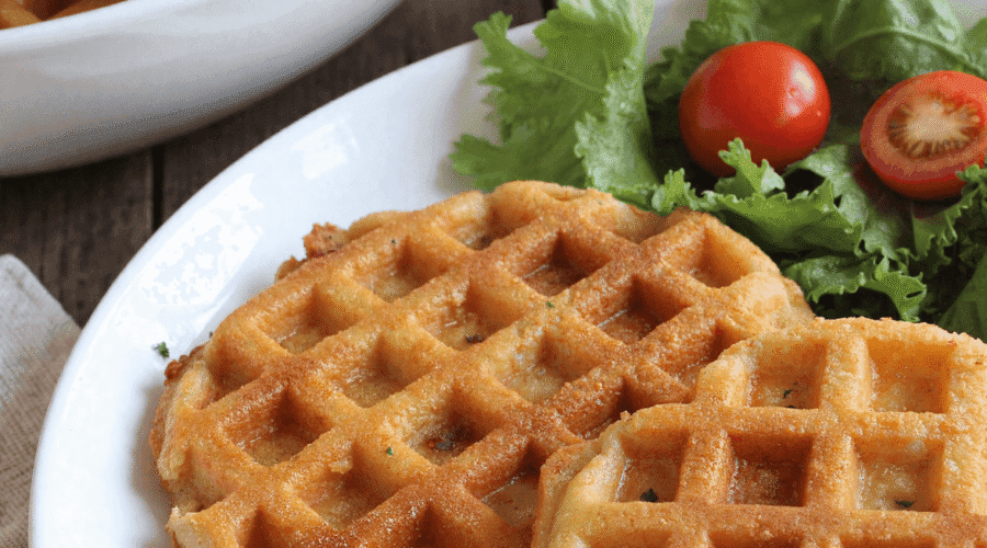 Plate of three golden crispy low carb hashbrown chaffles with a side of leafy greens and cherry tomatoes, perfect for healthy meal prep or low carb brunch.