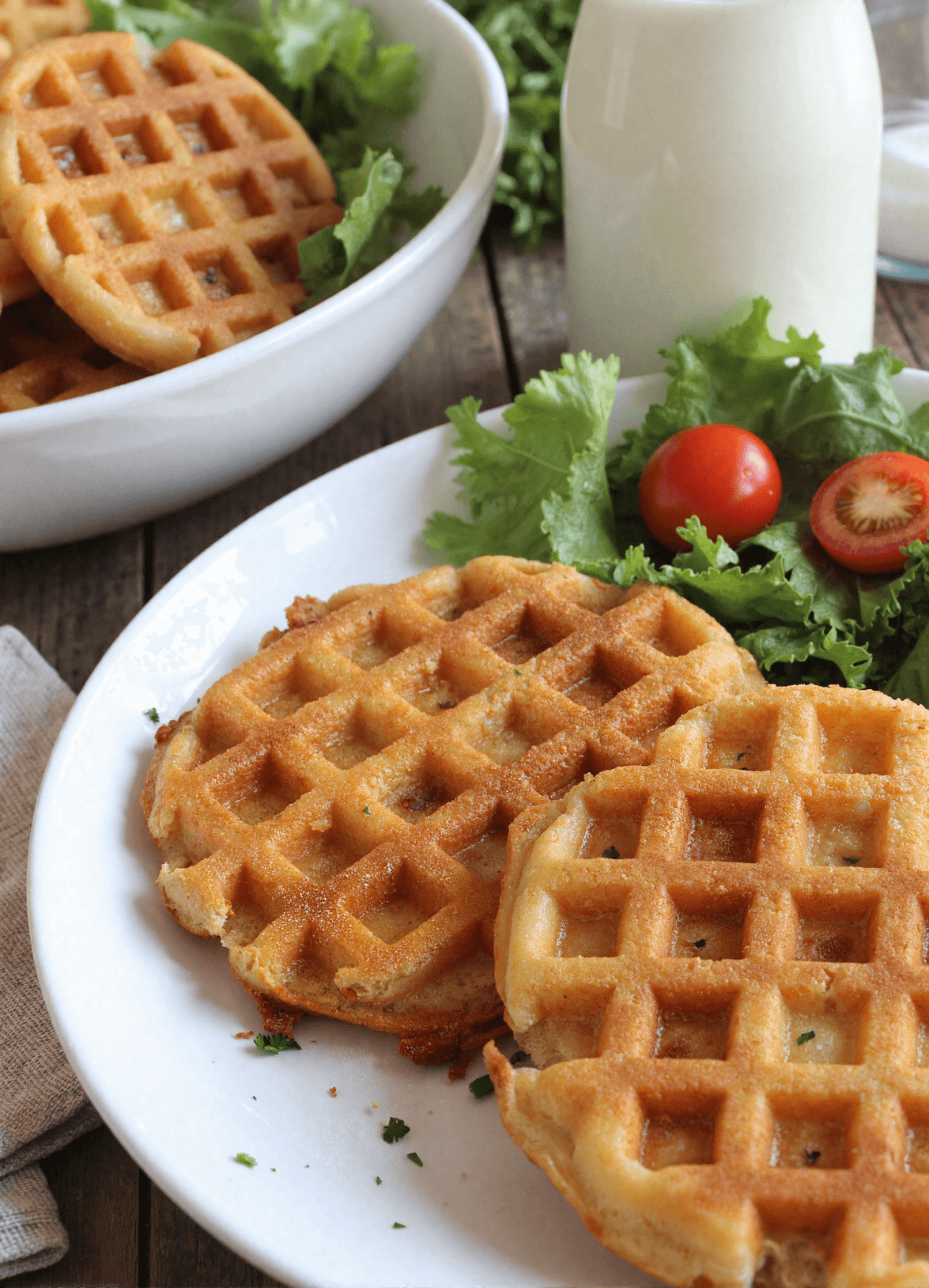 Plate of three golden crispy low carb hashbrown chaffles with a side of leafy greens and cherry tomatoes, perfect for healthy meal prep or low carb brunch.