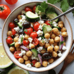 Bowl of Mediterranean chickpea salad with chickpeas, cucumber, cherry tomatoes, kalamata olives, red onion, parsley, and crumbled feta, served with lemon slices.