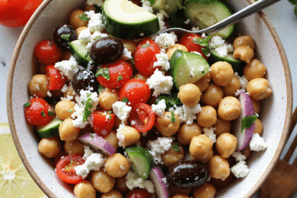 Bowl of Mediterranean chickpea salad with chickpeas, cucumber, cherry tomatoes, kalamata olives, red onion, parsley, and crumbled feta, served with lemon slices.