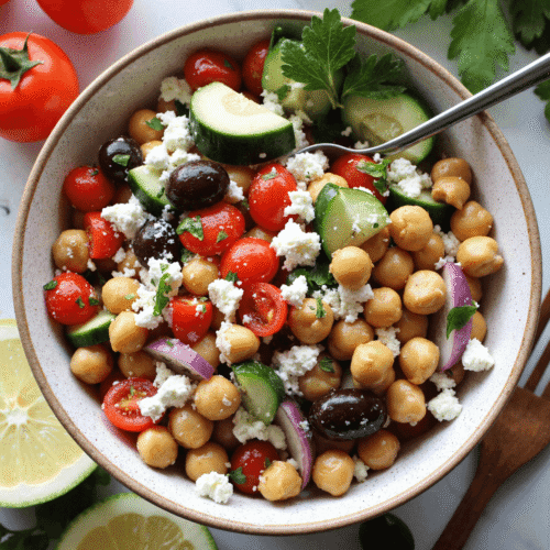 Bowl of Mediterranean chickpea salad with chickpeas, cucumber, cherry tomatoes, kalamata olives, red onion, parsley, and crumbled feta, served with lemon slices.