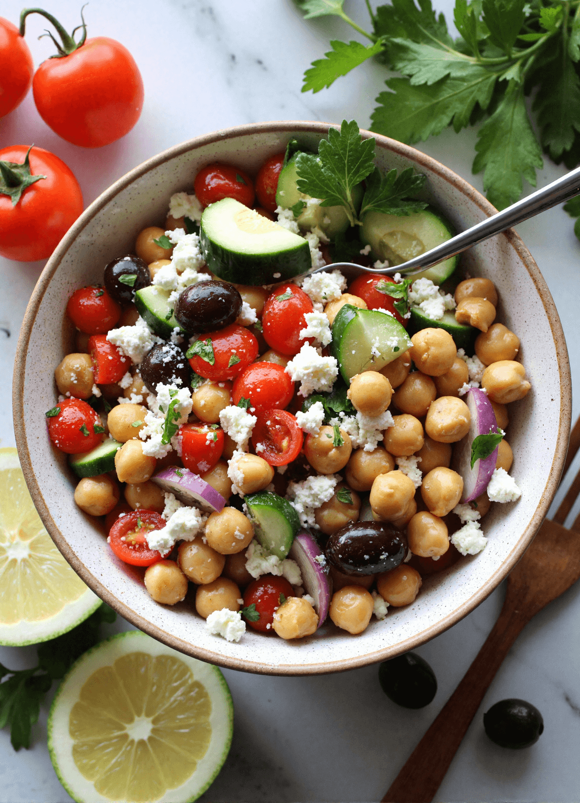 Bowl of Mediterranean chickpea salad with chickpeas, cucumber, cherry tomatoes, kalamata olives, red onion, parsley, and crumbled feta, served with lemon slices.