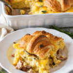 Serving of overnight croissant breakfast casserole with eggs, ham, and cheese on a white plate, garnished with fresh herbs and croissants baked on top.