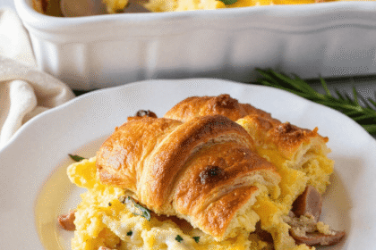 Serving of overnight croissant breakfast casserole with eggs, ham, and cheese on a white plate, garnished with fresh herbs and croissants baked on top.