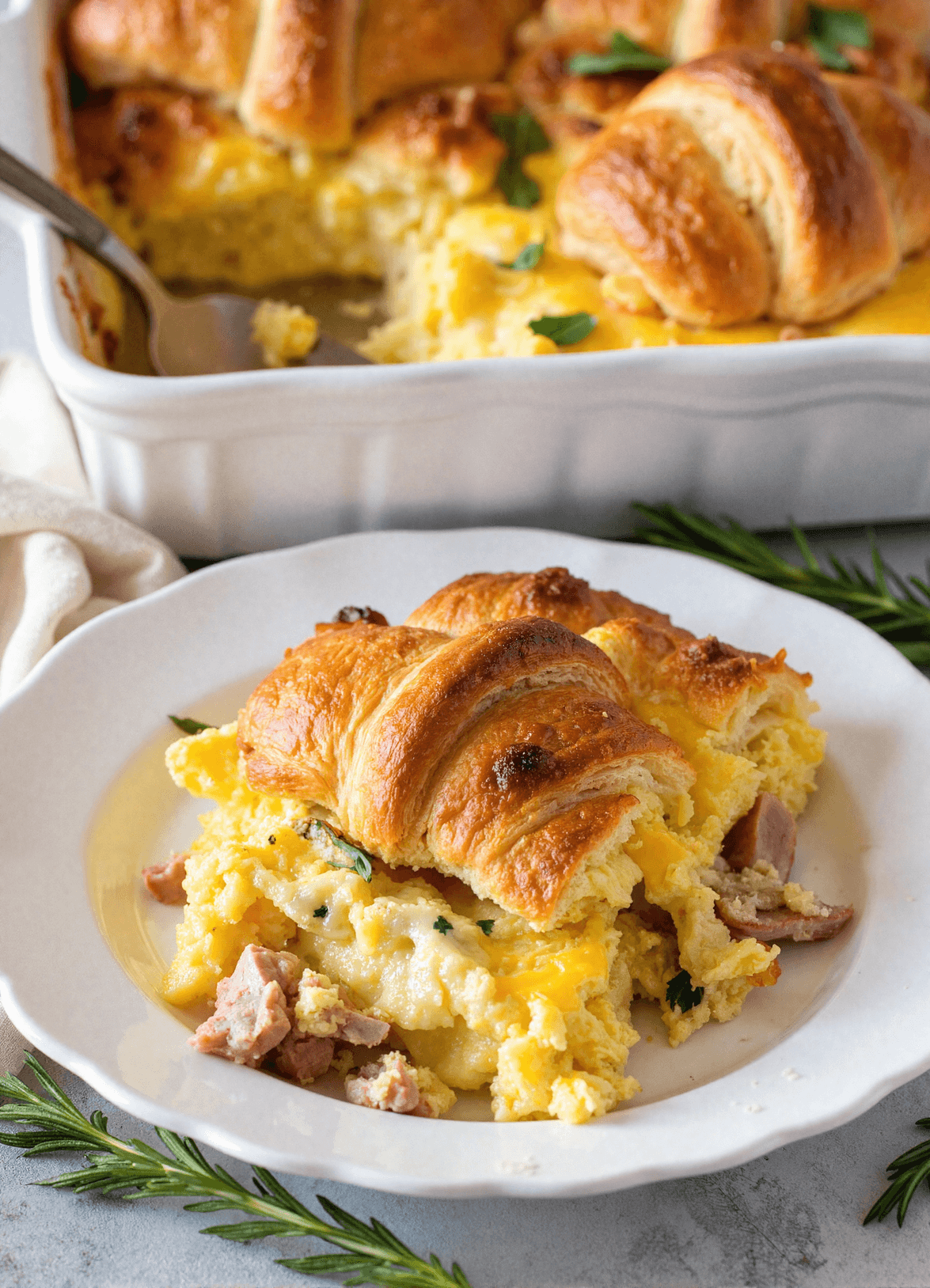 Serving of overnight croissant breakfast casserole with eggs, ham, and cheese on a white plate, garnished with fresh herbs and croissants baked on top.