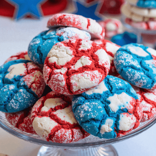 Red white and blue patriotic crinkle cookies stacked on a glass cake stand, dusted with powdered sugar for 4th of July celebrations.