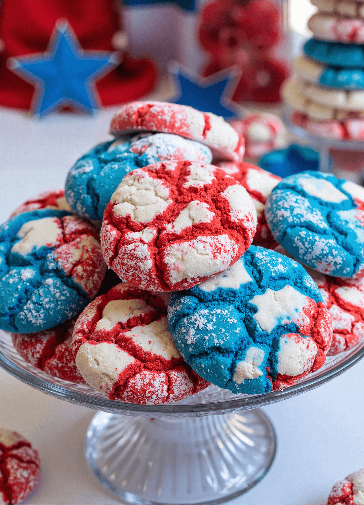 Red white and blue patriotic crinkle cookies stacked on a glass cake stand, dusted with powdered sugar for 4th of July celebrations.
