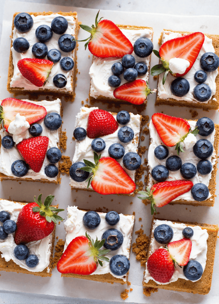 Close-up of red, white, and blue cheesecake bars decorated with whipped cream, strawberries, blueberries, and blue star-shaped toppings.