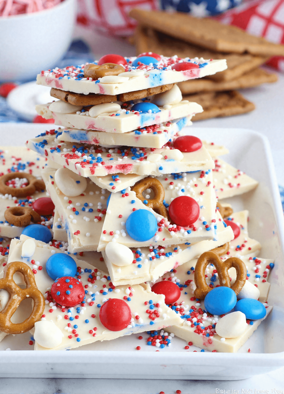 Stacked pieces of red white and blue pretzel bark made with white chocolate, patriotic sprinkles, mini pretzels, and M&Ms on a marble tray.
