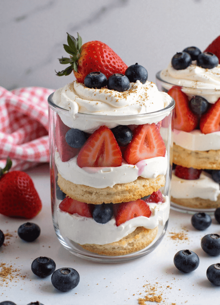 Close-up of a layered berry shortcake trifle in a glass with fresh strawberries, blueberries, whipped cream, and biscuit rounds.