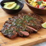 Sliced grilled steak topped with chimichurri sauce on a wooden board with avocado and corn salad in the background