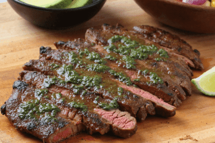 Sliced grilled steak topped with chimichurri sauce on a wooden board with avocado and corn salad in the background