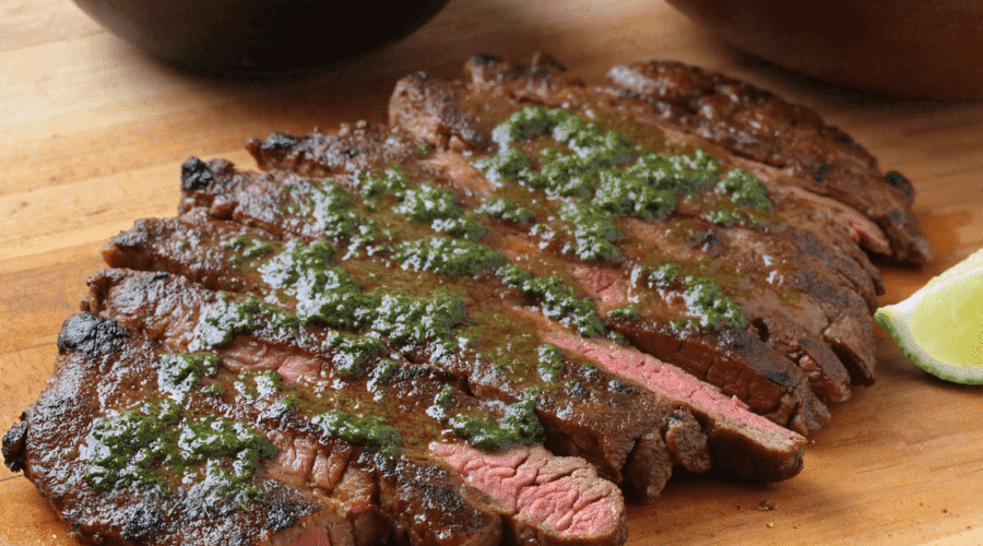Sliced grilled steak topped with chimichurri sauce on a wooden board with avocado and corn salad in the background