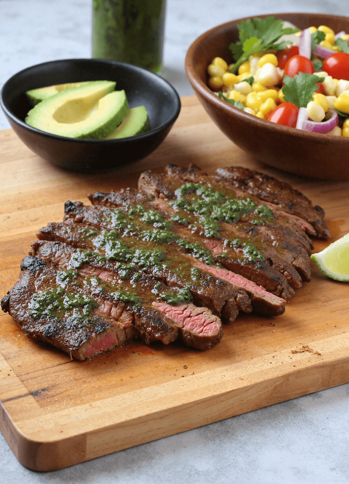 Sliced grilled steak topped with chimichurri sauce on a wooden board with avocado and corn salad in the background