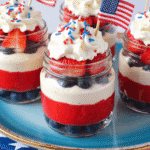 Fourth of July Cheesecake Cups in mason jars layered with blueberries, strawberry puree, cream cheese filling, and whipped cream, topped with red, white, and blue sprinkles and tiny American flags.