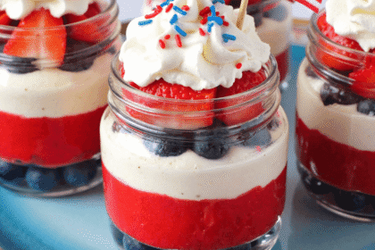 Fourth of July Cheesecake Cups in mason jars layered with blueberries, strawberry puree, cream cheese filling, and whipped cream, topped with red, white, and blue sprinkles and tiny American flags.
