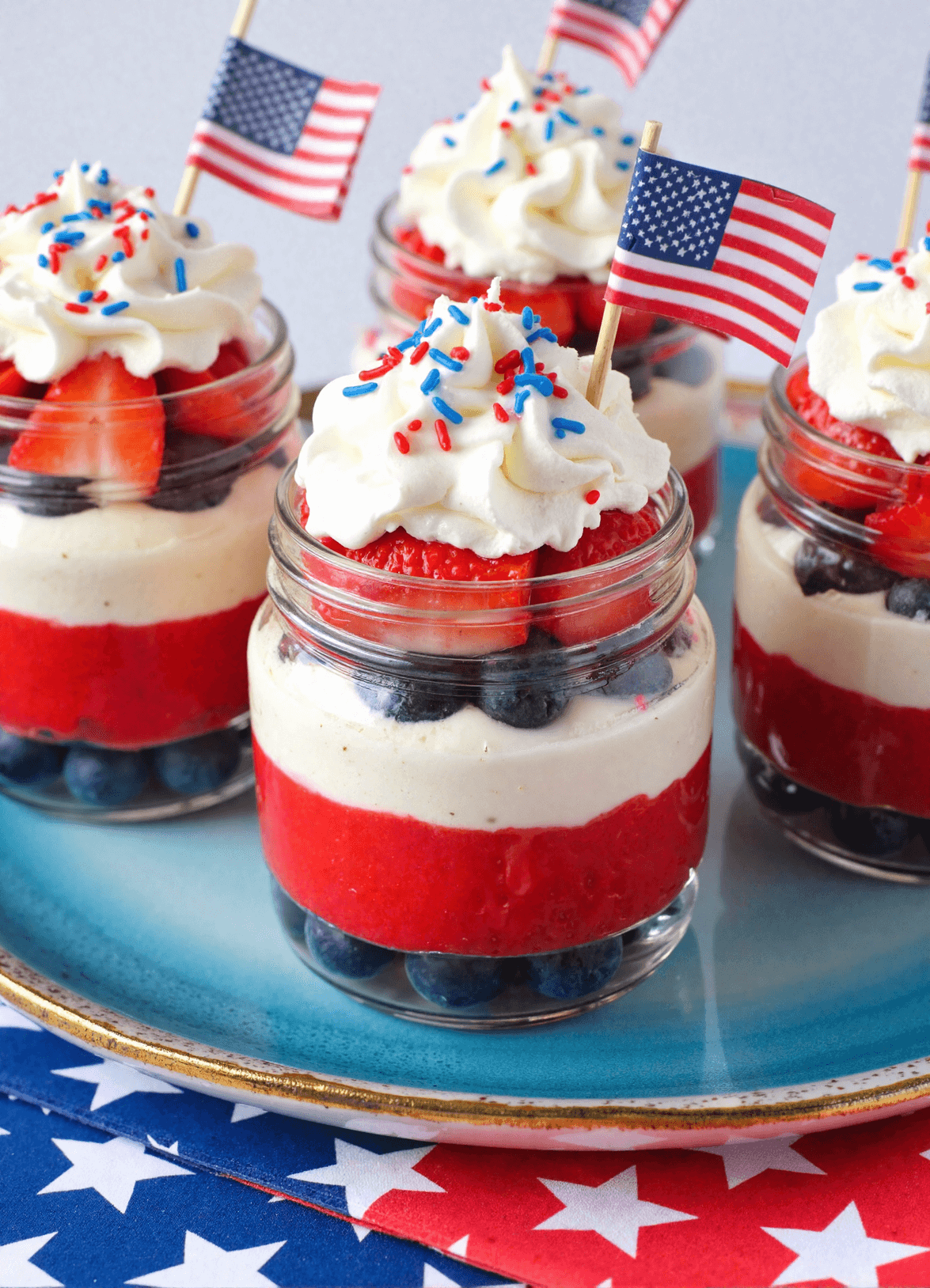 Fourth of July Cheesecake Cups in mason jars layered with blueberries, strawberry puree, cream cheese filling, and whipped cream, topped with red, white, and blue sprinkles and tiny American flags.