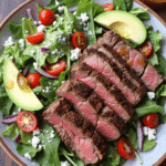 grilled steak salad with avocado, cherry tomatoes, feta cheese, and baby greens on a ceramic plate