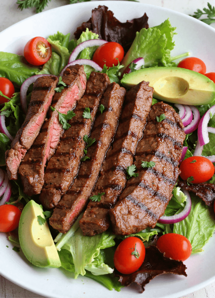 grilled sirloin steak salad with avocado slices, grape tomatoes, red onion, and mixed greens on a white plate