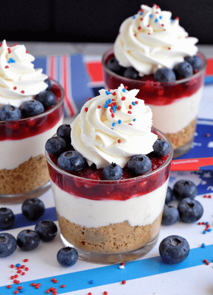 No-bake blueberry cheesecake cups with graham cracker crust, vanilla cream cheese filling, berry topping, and whipped cream swirls decorated with red and blue sprinkles.