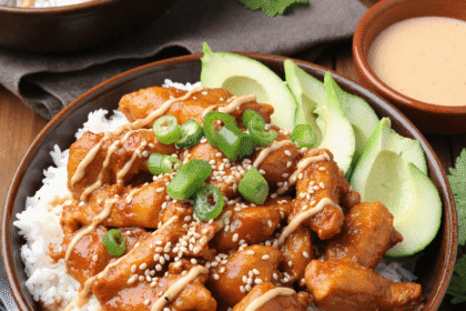 Spicy honey-ginger chicken over white rice, topped with yum yum sauce, sliced avocado, sesame seeds, and green onions in a brown bowl.