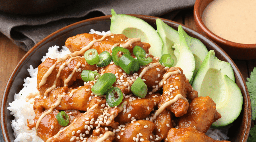 Spicy honey-ginger chicken over white rice, topped with yum yum sauce, sliced avocado, sesame seeds, and green onions in a brown bowl.
