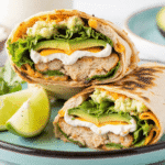 Toasted chicken avocado ranch burrito cut in half with layers of grilled chicken, cheddar, avocado, lettuce, and ranch, served with lime wedges on a turquoise plate.