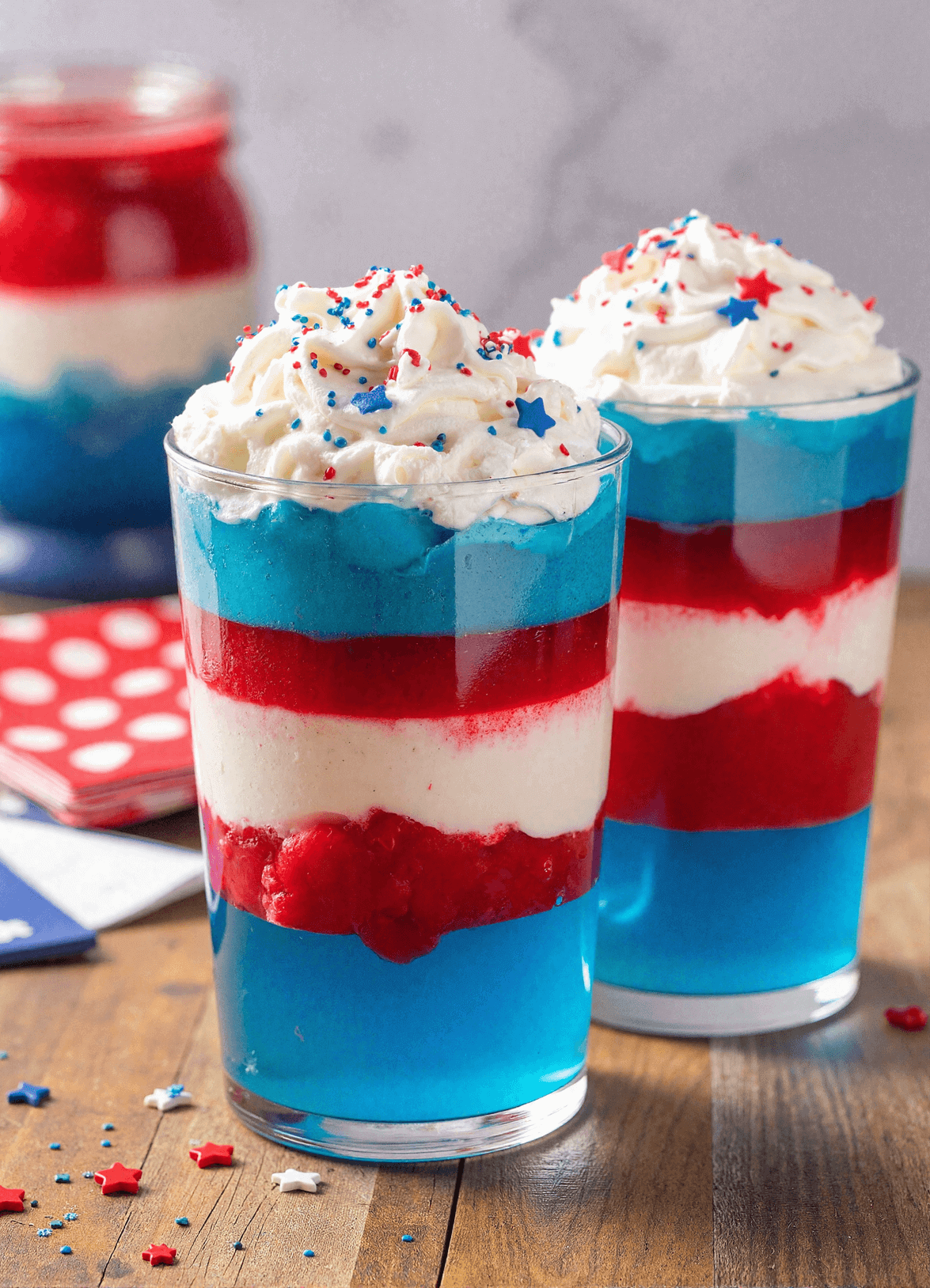 Fourth of July layered jello cups in red, white, and blue, topped with whipped cream and red-blue sprinkles.