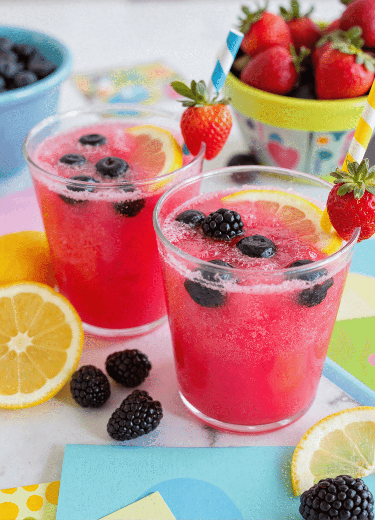 Two glasses of berry lemonade slush punch with lemon slices, blackberries, and blueberries on a colorful summer-themed table.