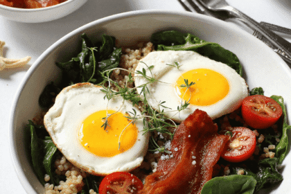 BLT breakfast bowl with fried eggs, crispy bacon, sautéed spinach, cherry tomatoes, and cooked grains served in a white bowl.
