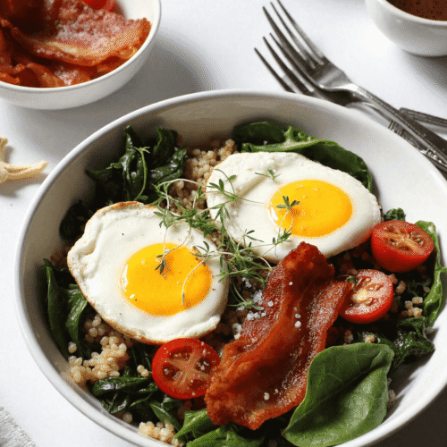 BLT breakfast bowl with fried eggs, crispy bacon, sautéed spinach, cherry tomatoes, and cooked grains served in a white bowl.