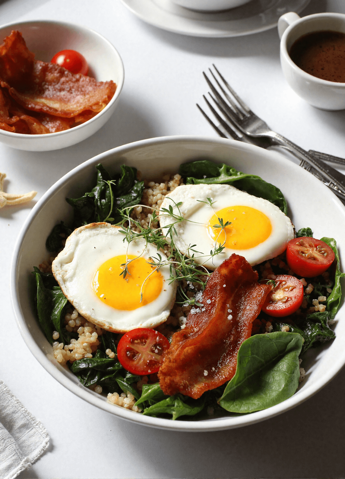 BLT breakfast bowl with fried eggs, crispy bacon, sautéed spinach, cherry tomatoes, and cooked grains served in a white bowl.