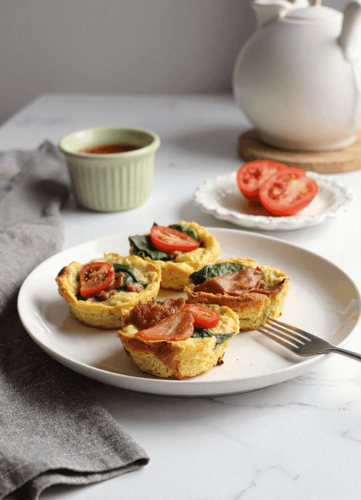 Four BLT frittata cups on a white plate, filled with crispy bacon, spinach, and cherry tomatoes, served on a marble counter with a side of sauce and fresh tomato slices.