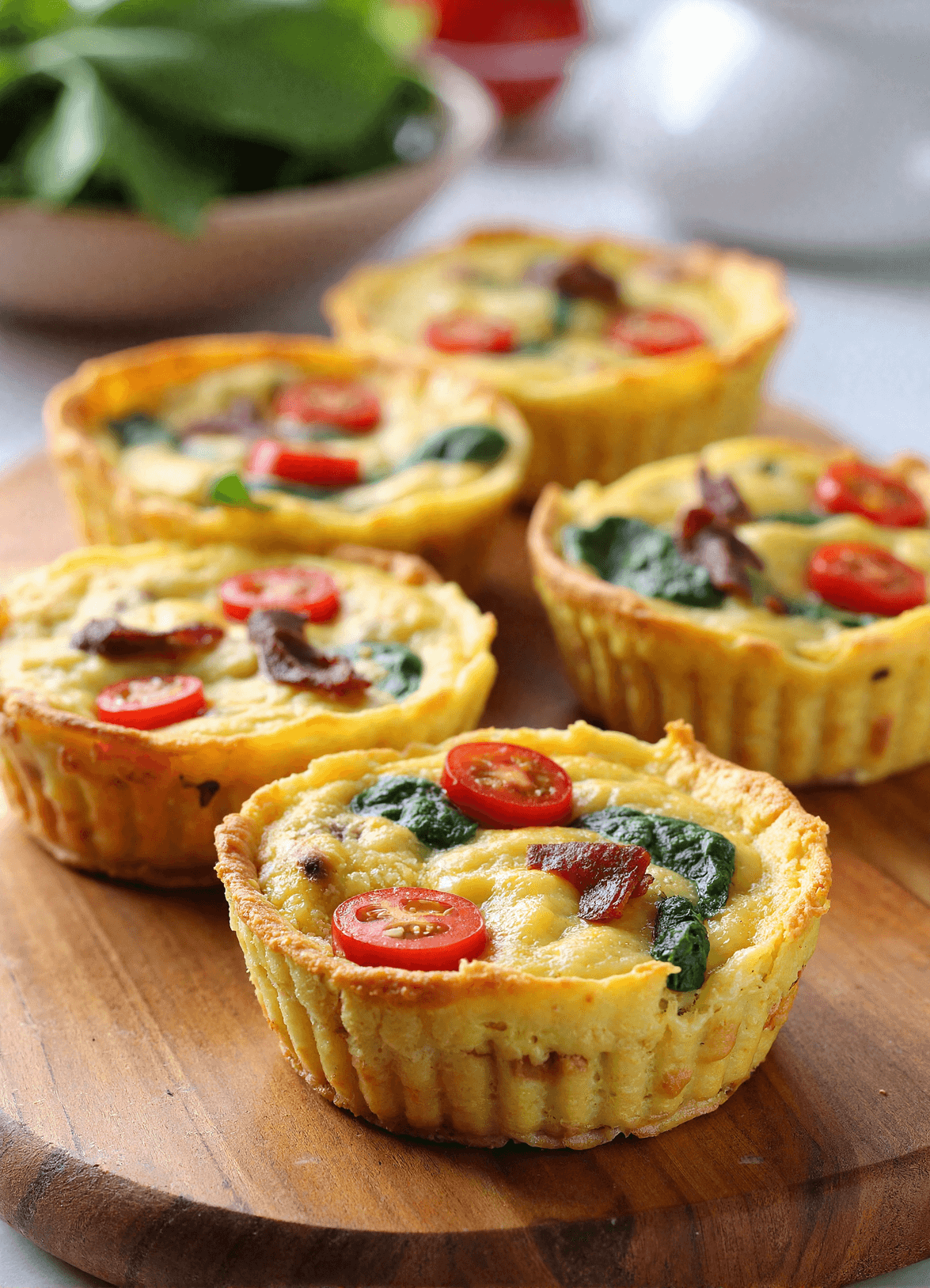 Close-up of mini BLT frittata muffins on a wooden board topped with spinach, tomatoes, and bacon pieces.