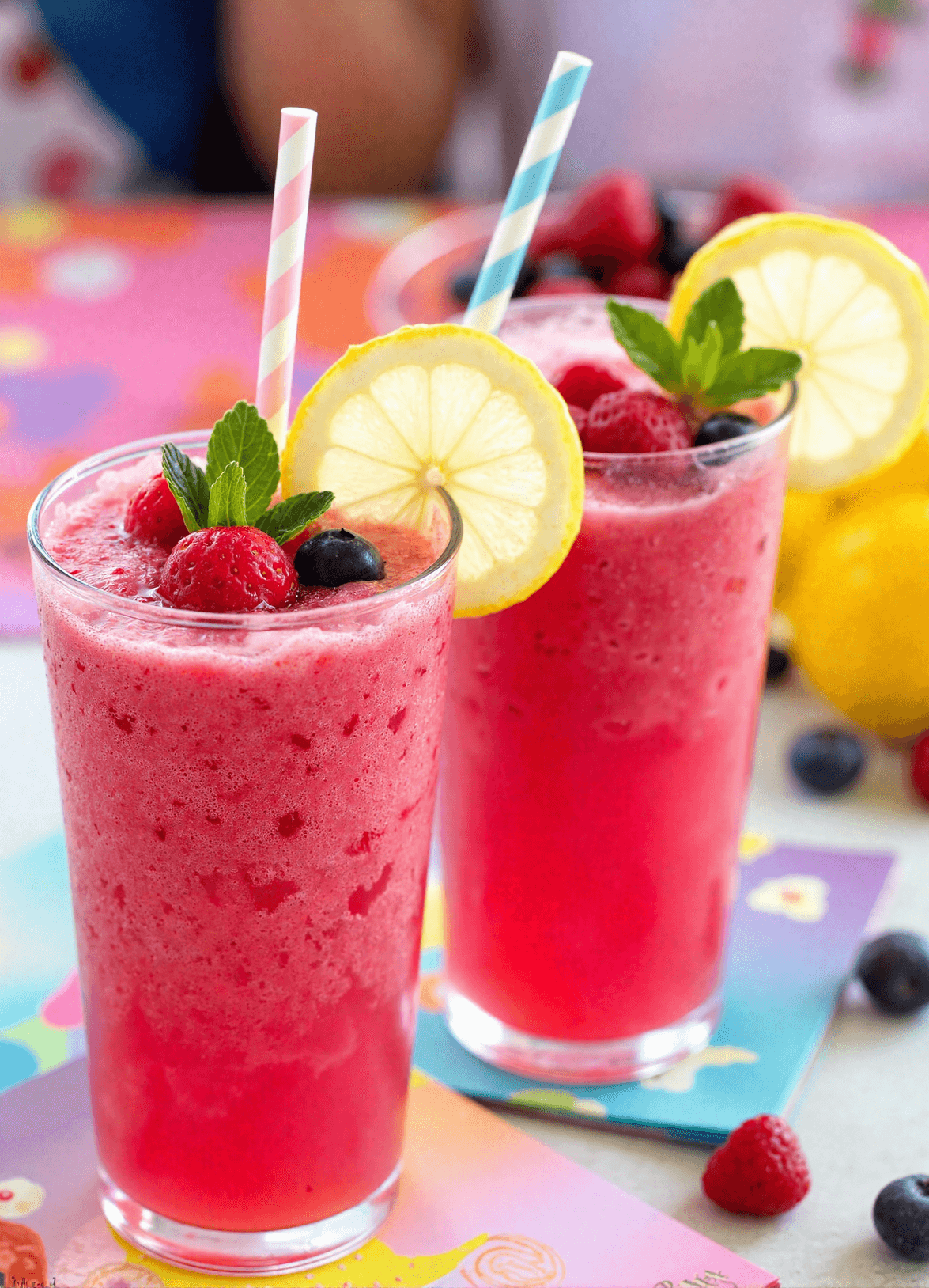Tall glasses of frozen berry lemonade punch topped with raspberries, blueberries, lemon wheels, and fresh mint on a bright table.