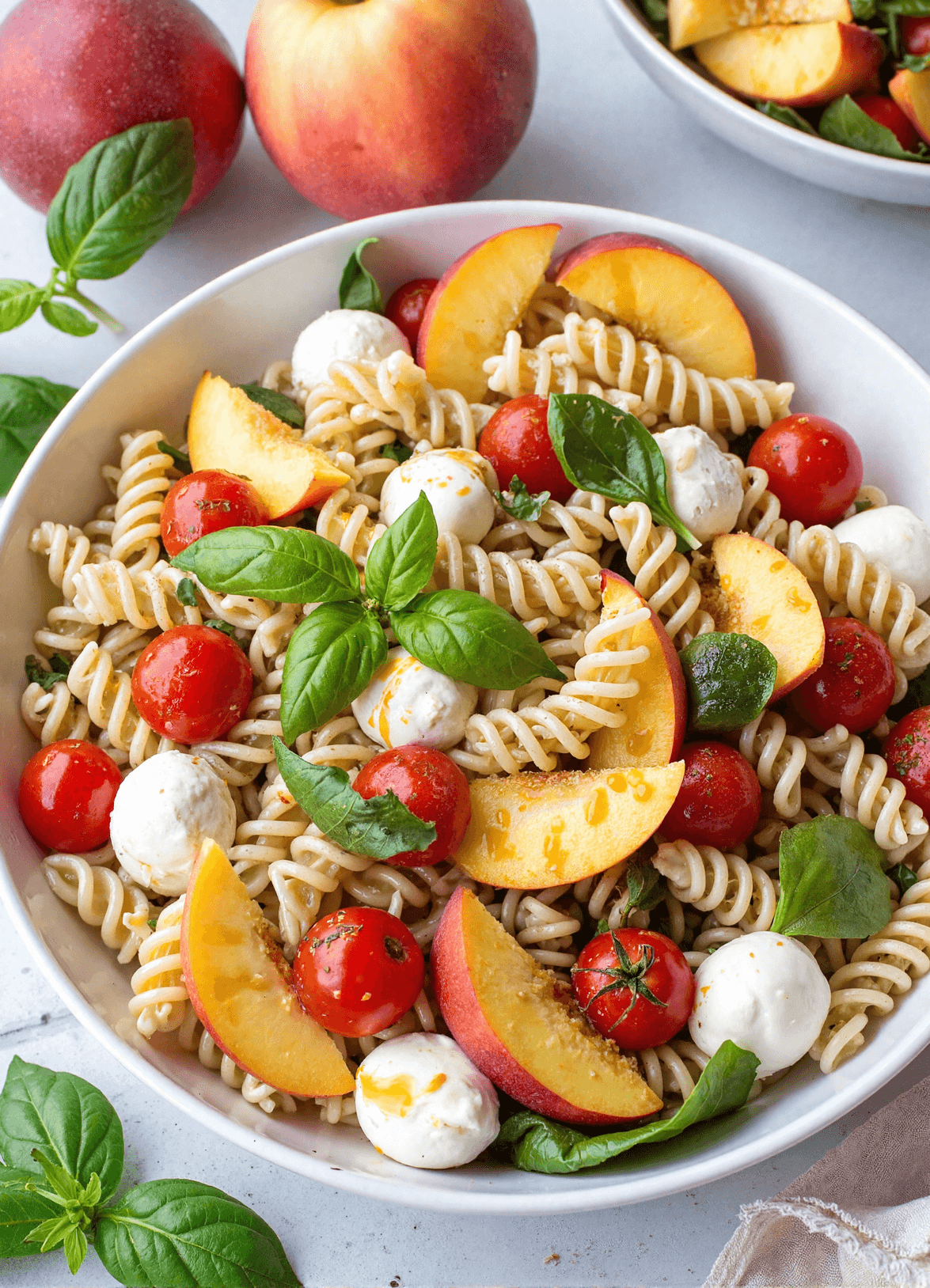 Overhead view of fruit Caprese pasta salad with sliced peaches, cherry tomatoes, mozzarella pearls, and fresh basil in a white serving bowl.