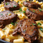 Seared garlic butter steak tips on cheesy rigatoni pasta, garnished with fresh parsley in a black skillet.