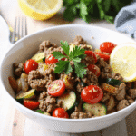 Close-up angled view of a white bowl filled with seasoned ground beef, zucchini, tomatoes, and bell peppers garnished with parsley and lemon—ideal for a low-carb Mediterranean dinner.