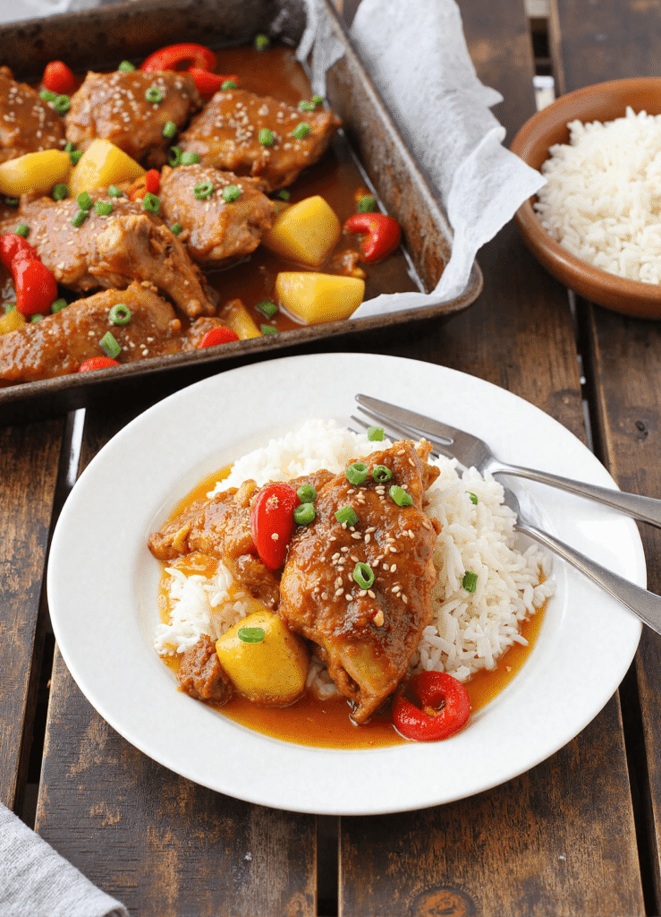 Hawaiian chicken thighs served over rice with pineapple, red peppers, and sesame seeds on a white plate beside a baking pan