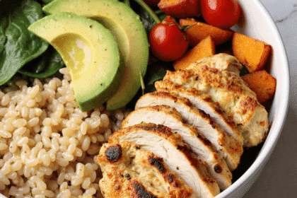 Sliced grilled chicken served over quinoa with spinach, avocado, roasted sweet potatoes, and cherry tomatoes in a white bowl.