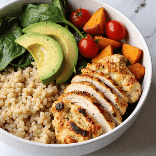 Sliced grilled chicken served over quinoa with spinach, avocado, roasted sweet potatoes, and cherry tomatoes in a white bowl.