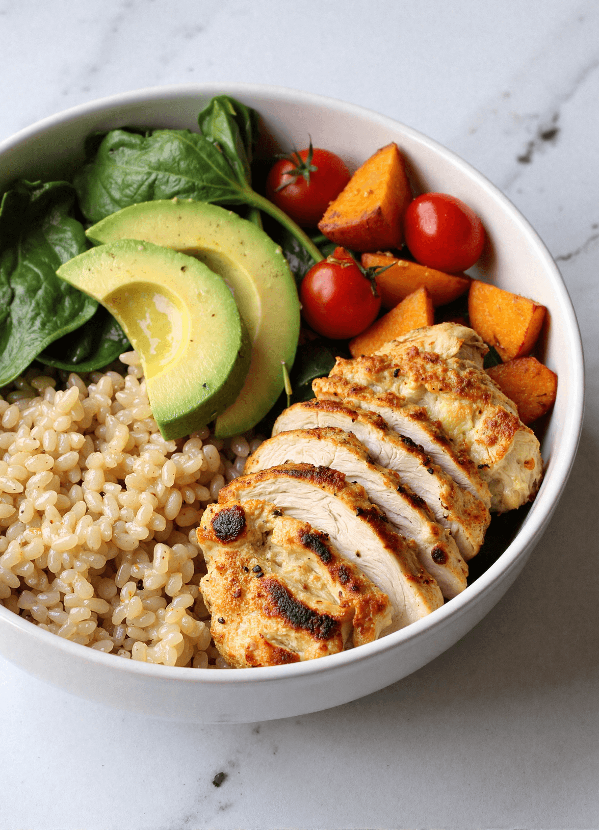 Sliced grilled chicken served over quinoa with spinach, avocado, roasted sweet potatoes, and cherry tomatoes in a white bowl.