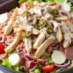 Close-up of Italian grinder salad with sliced turkey, salami, hearts of palm, cherry tomatoes, and creamy dressing in a white bowl on a wooden surface.