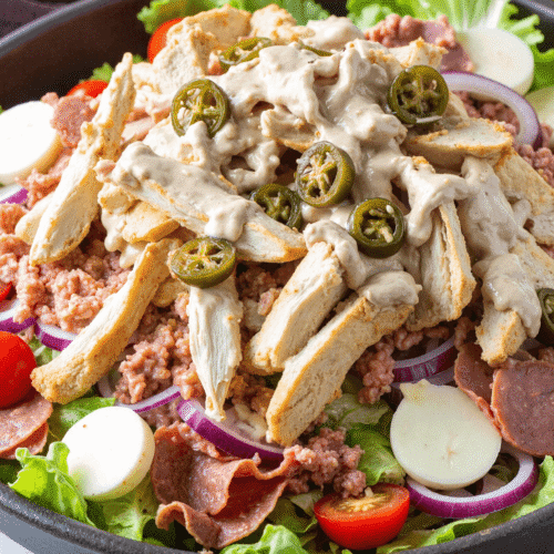 Close-up of Italian grinder salad with sliced turkey, salami, hearts of palm, cherry tomatoes, and creamy dressing in a white bowl on a wooden surface.