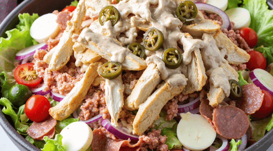 Close-up of Italian grinder salad with sliced turkey, salami, hearts of palm, cherry tomatoes, and creamy dressing in a white bowl on a wooden surface.